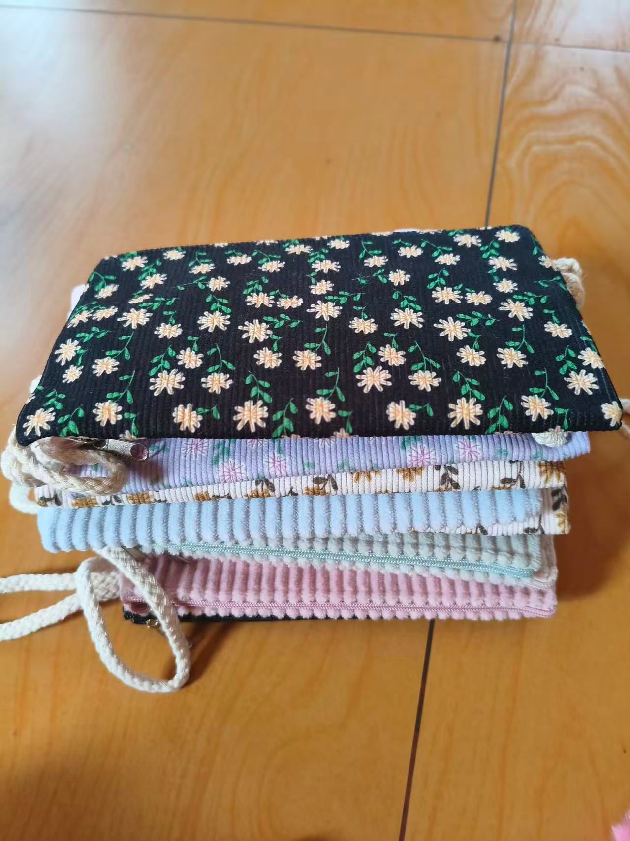 A neat stack of folded corduroy zip pouches in floral and plain patterns, with woven drawstrings, resting on a wooden floor.